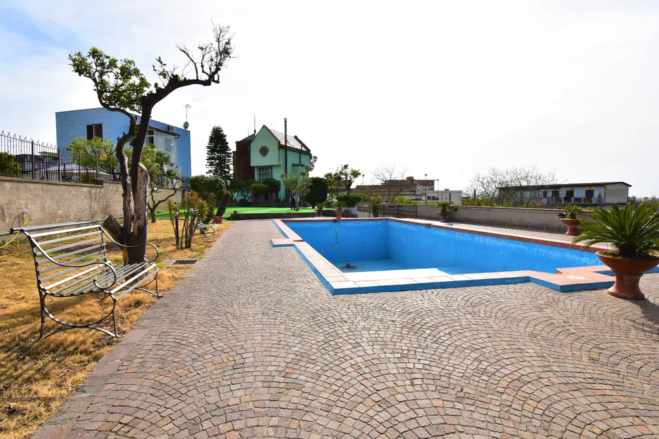 La piscina vuota e il vialetto in porfido nel giardino di una villa, con una seconda villa sullo sfondo
